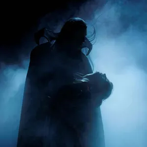 Zwei Figuren in einer d&uuml;steren B&uuml;hnenszene: Eine dunkel gekleidete Gestalt h&auml;lt eine weitere Person im Arm. Umgeben von blauem Nebel entsteht die charakteristische, geheimnisvolle Atmosph&auml;re von &sbquo;Tanz der Vampire