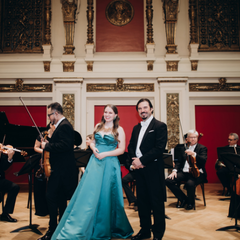 Kammerorchester auf einer Konzertb&uuml;hne in einem prunkvollen Saal. In der Mitte stehen drei Personen in Abendgarderobe vor den Musikerinnen und Musikern mit Streichinstrumenten und Fl&uuml;gel