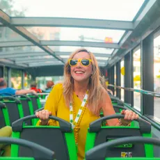 A person wearing yellow sunglasses and yellow clothing is sitting on the upper deck of a sightseeing bus with a glass roof. The green-and-black seats and the bright daylight convey the experience of a city tour.