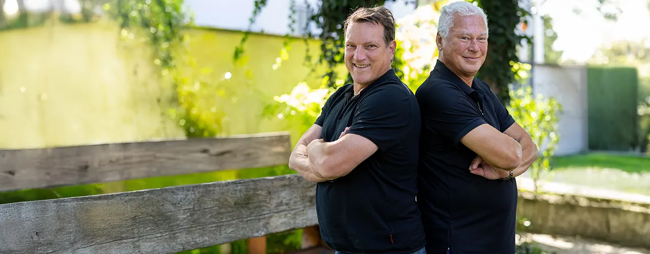 Zwei Personen in dunklen Poloshirts stehen mit verschr&auml;nkten Armen R&uuml;cken an R&uuml;cken vor einem Holzgel&auml;nder. Im Hintergrund sind B&auml;ume und eine sonnige Gr&uuml;nanlage zu sehen.