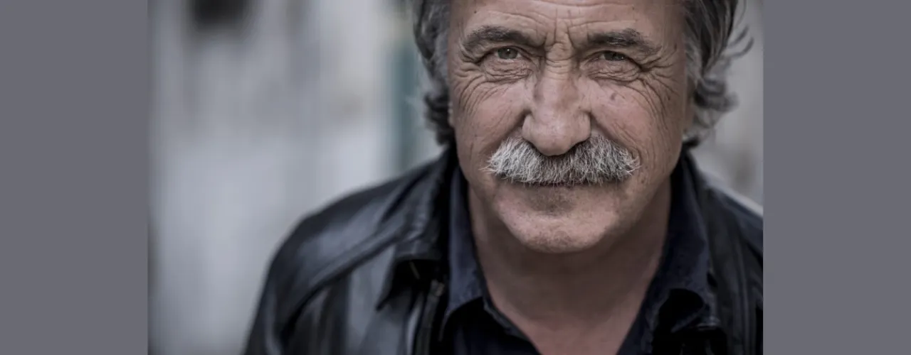 A close-up of a person with a distinctive mustache and a leather jacket. The face fills the right side of the frame, and the person is looking directly at the camera. The background is out of focus and rendered in muted shades of gray.