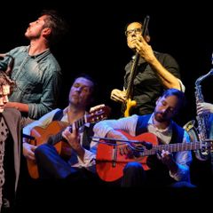 Mehrere Musikerinnen und Musiker sowie eine tanzende Person stehen gemeinsam auf einer dunkel beleuchteten B&uuml;hne. Gitarren, Saxofon, Bass und Percussion pr&auml;gen die Konzertszene mit Fokus auf Flamenco- und Live-Musik.