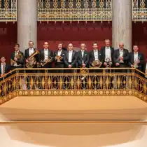 Gruppe von Musikerinnen und Musikern mit Blechblasinstrumenten auf einem verzierten Balkon in einem Konzertsaal. Goldfarbene Gel&auml;nder, rote Wandfl&auml;chen und hohe S&auml;ulen pr&auml;gen die symmetrische Innenarchitektur