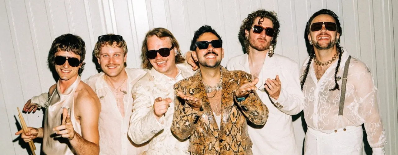 Die Band Roy Bianco &amp; Abbrunzati Boys posiert gemeinsam vor einer hellen Wand. Sechs Bandmitglieder tragen auff&auml;llige B&uuml;hnenoutfits in Creme-, Wei&szlig;- und Goldt&ouml;nen sowie Sonnenbrillen; im Mittelpunkt steht ein Anzug mit Schlangenmuster.
