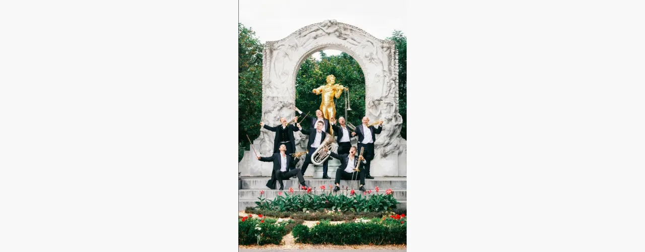 Sechs Musiker mit Blechblasinstrumenten posieren vor einem kunstvollen wei&szlig;en Pavillon mit goldener Statue in einer Parkanlage. Die Gruppe ist auf mehreren Stufen arrangiert, umgeben von Blumenbeeten und Gr&uuml;nfl&auml;chen
