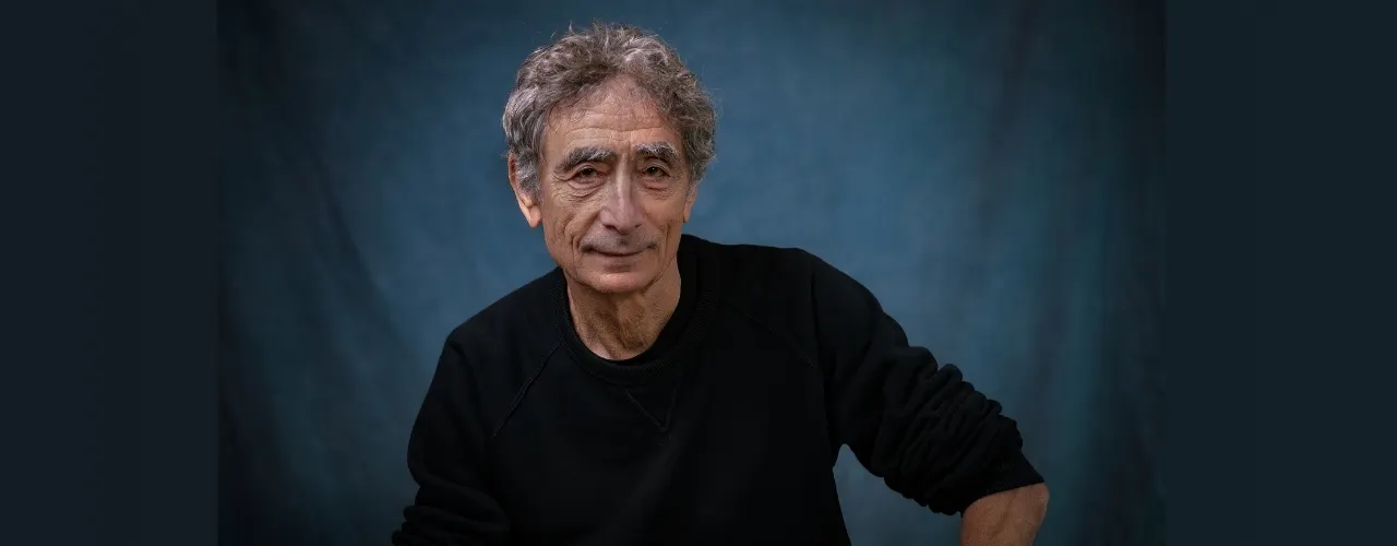 A portrait of a person with gray, curly hair wearing a dark sweater, sitting in front of a blue-lit background. The person is looking directly at the camera; their upper body and face are clearly visible.