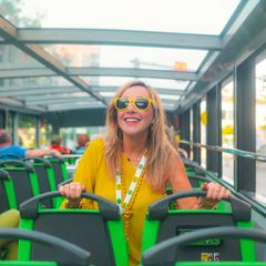 Person mit gelber Sonnenbrille und gelber Kleidung sitzt im Oberdeck eines Sightseeingbusses mit Glasdach. Die gr&uuml;n-schwarzen Sitze und das helle Tageslicht vermitteln das Erlebnis einer Stadtrundfahrt
