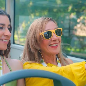 A person wearing yellow sunglasses and yellow clothing is sitting on the upper deck of a sightseeing bus with a glass roof. The green-and-black seats and the bright daylight convey the experience of a city tour.