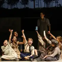 Several children are sitting and kneeling on a theater stage, raising their arms high. Another person is standing in the background. The scene is bathed in warm light, with a dark forest backdrop visible behind it.