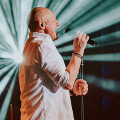 A person on a stage holding a microphone, with a bright, radial light effect in the background. The scene depicts a live performance with intense stage lighting and a concert atmosphere.