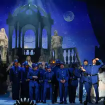 A stage scene featuring a group of performers in blue uniforms in front of a monumental staircase and sculptures. In the background, a starry night sky, moonlight, and a temple-like backdrop can be seen.