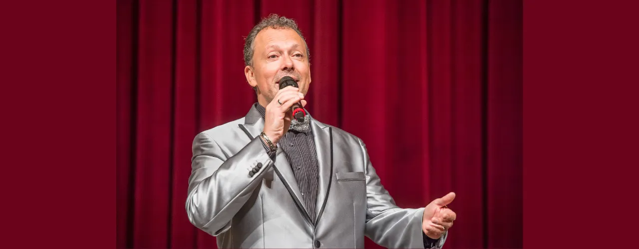 Promotional image for &ldquo;Life Is a Joke&rdquo;: Wolf Frank stands on stage with a microphone in front of a red theater curtain. Dressed in a light-colored suit, he presents his show in a classic cabaret and variety show atmosphere.