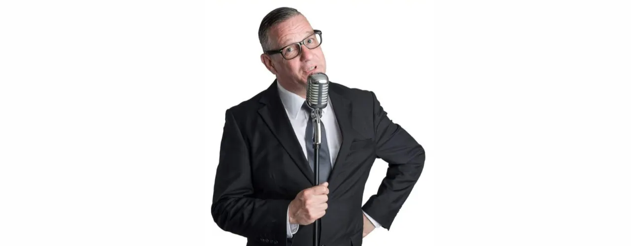 Manfred Distel steht im dunklen Anzug an einem klassischen Standmikrofon vor hellem Hintergrund.